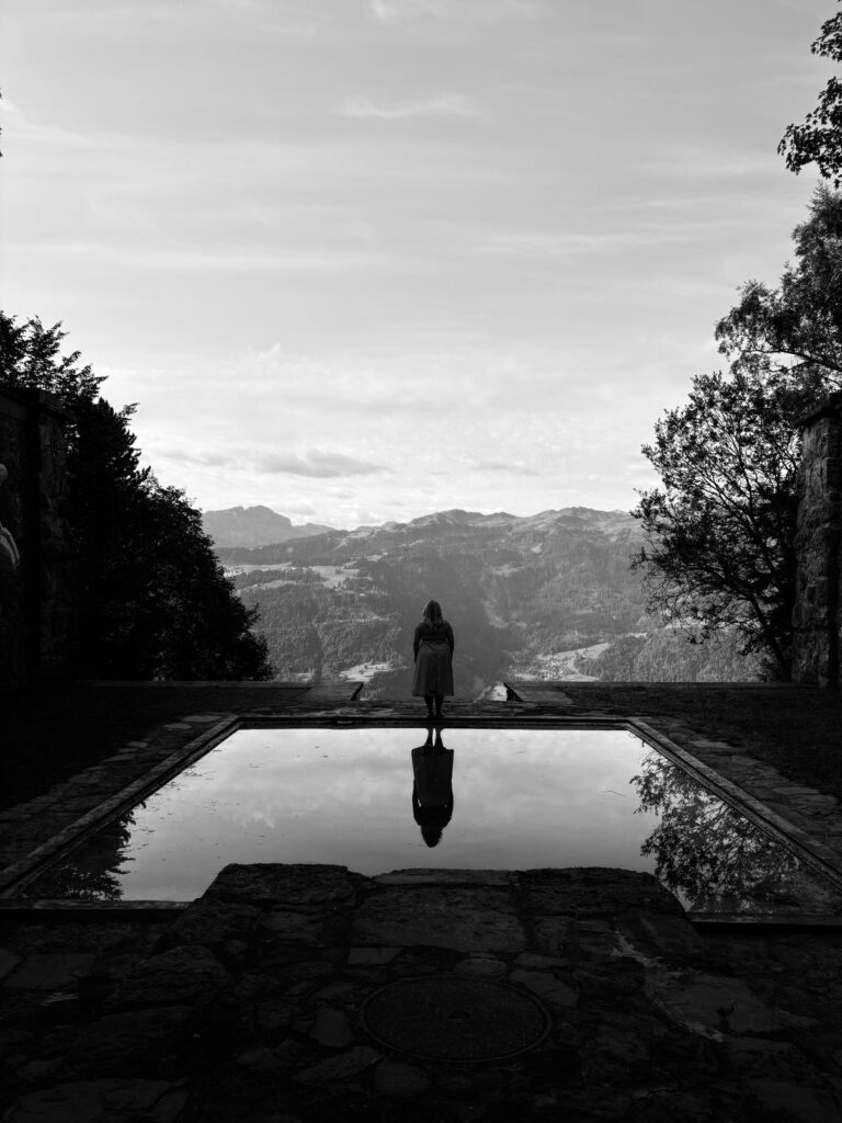 Frau steht auf einem höher gelegenenen Ort, hinter hier eine spiegelung ihrer Silouette im Wasser vor ihr eine Bergkulisse